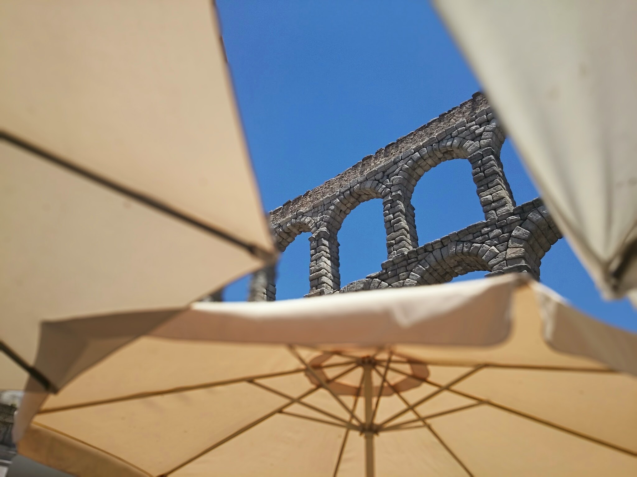 Fragment starożytnego akweduktu z kamienia, widziany spod beżowego parasola przeciwsłonecznego na tle błękitnego nieba.
