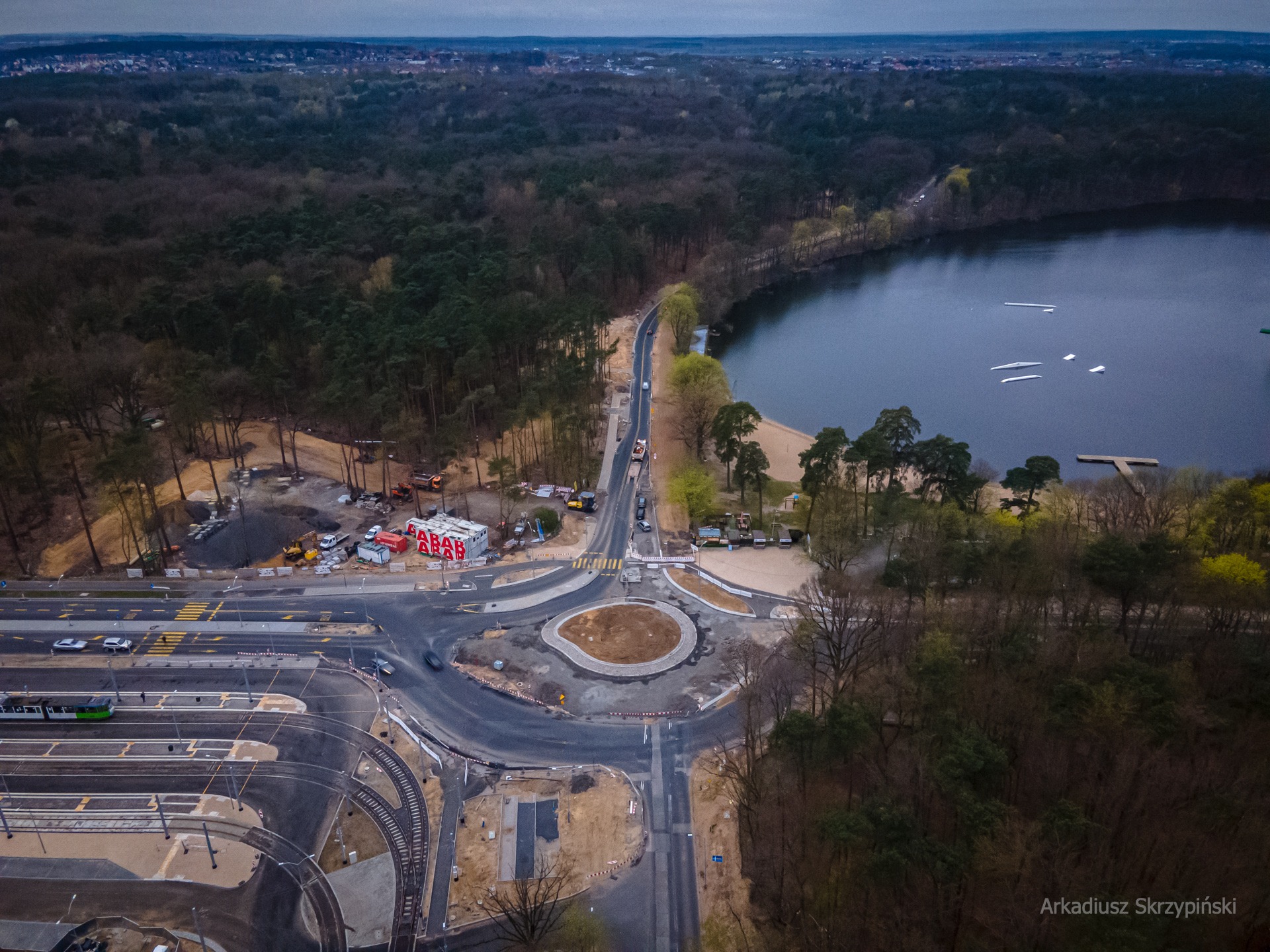 Zdjęcie z drona przedstawia skrzyżowanie z rondem w trakcie budowy, obok torów tramwajowych i jeziora z wake parkiem, otoczone lasem.