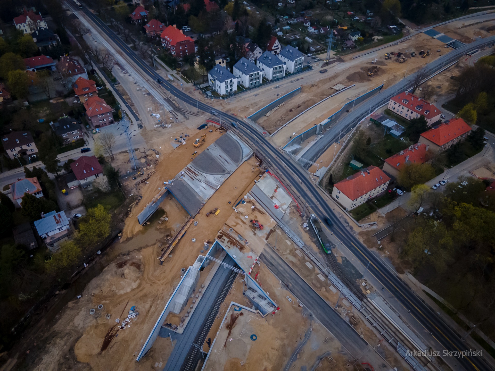 Widok z lotu ptaka na budowę wiaduktu kolejowego w otoczeniu domów jednorodzinnych i bloków mieszkalnych, z widocznymi maszynami budowlanymi i fragmentem przejeżdżającego pociągu.