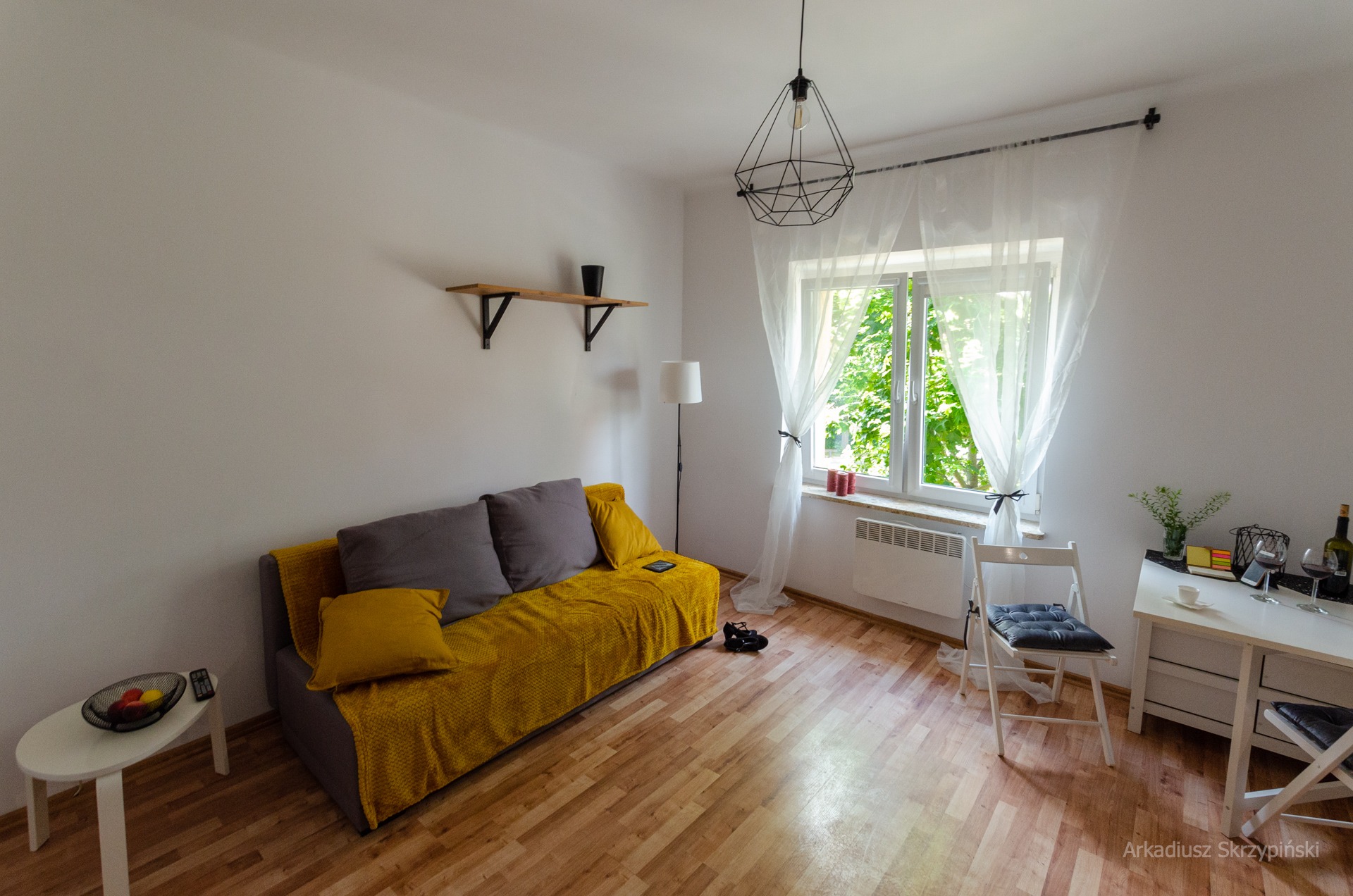 Jasne, minimalistyczne wnętrze pokoju dziennego z żółtą sofą, białym biurkiem i geometryczną lampą wiszącą, widok na zielone drzewa za oknem.