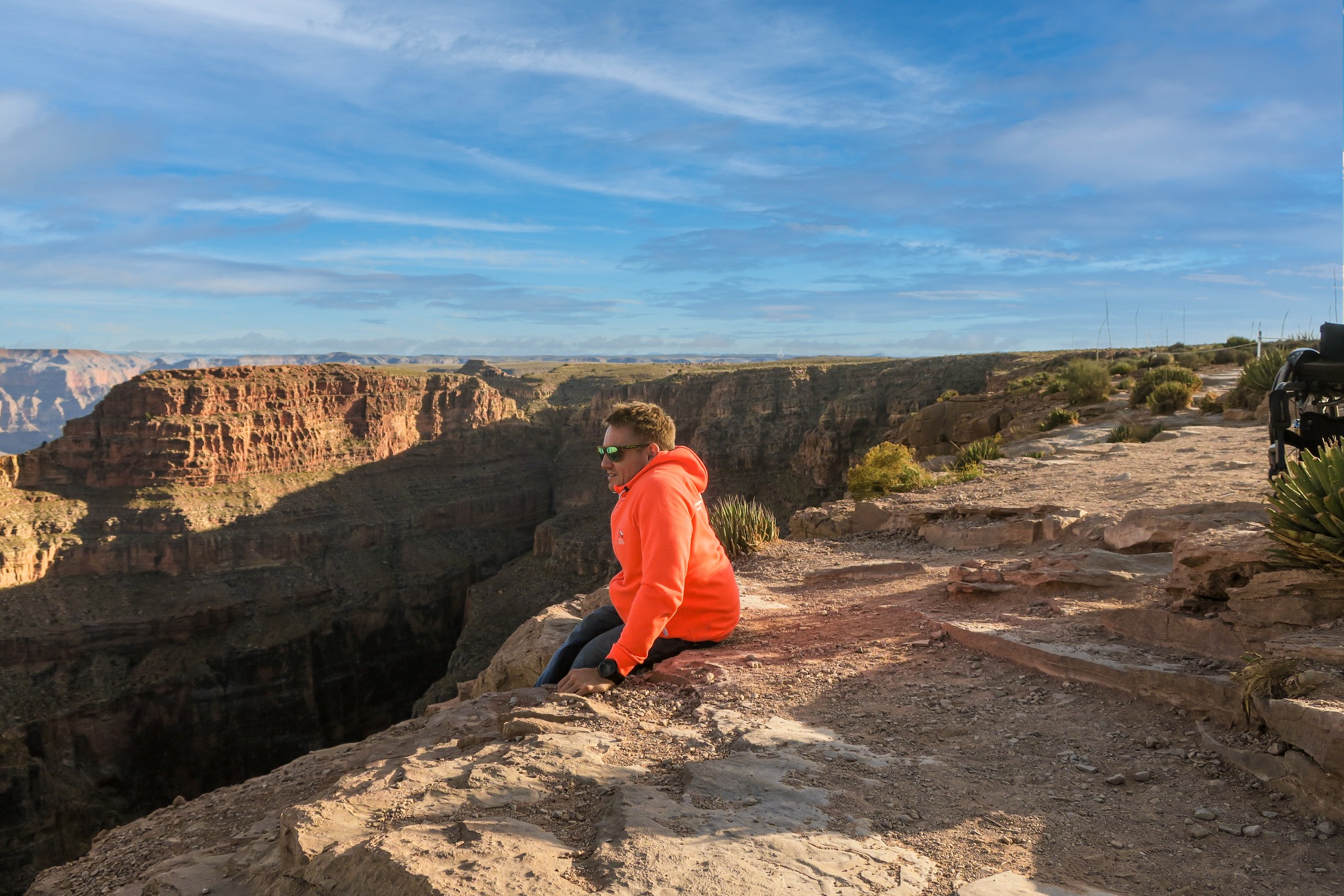 Mężczyzna w pomarańczowej bluzie siedzi na krawędzi skalnego klifu z widokiem na rozległy kanion i błękitne niebo.