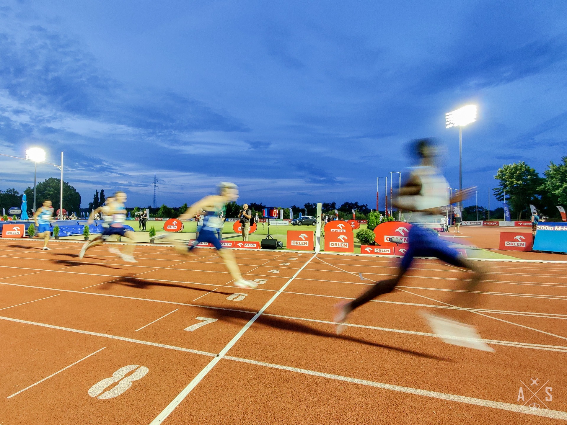 Rozmazane ujęcie biegnących sportowców na bieżni lekkoatletycznej, w tle reklamy Orlen, oświetlenie stadionowe i zachmurzone niebo.