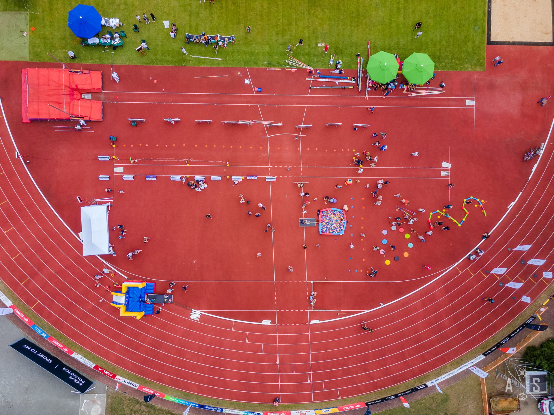 Widok z lotu ptaka na stadion lekkoatletyczny w Szczecinie podczas zawodów. Czerwona bieżnia z białymi liniami, liczne osoby, stanowiska z parasolami, skocznia wzwyż i dmuchany zamek.