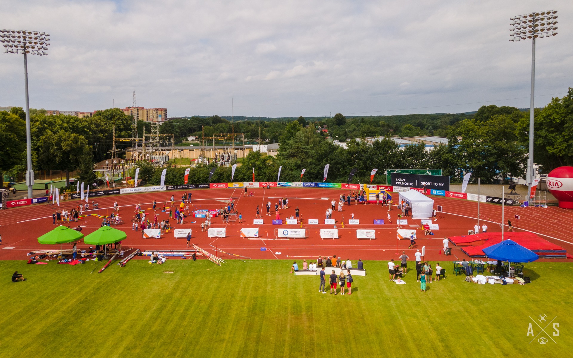 Widok z góry na stadion lekkoatletyczny z licznymi uczestnikami i kibicami podczas zawodów skoku o tyczce, z widocznymi stanowiskami sędziowskimi, namiotami i flagami sponsorskimi.