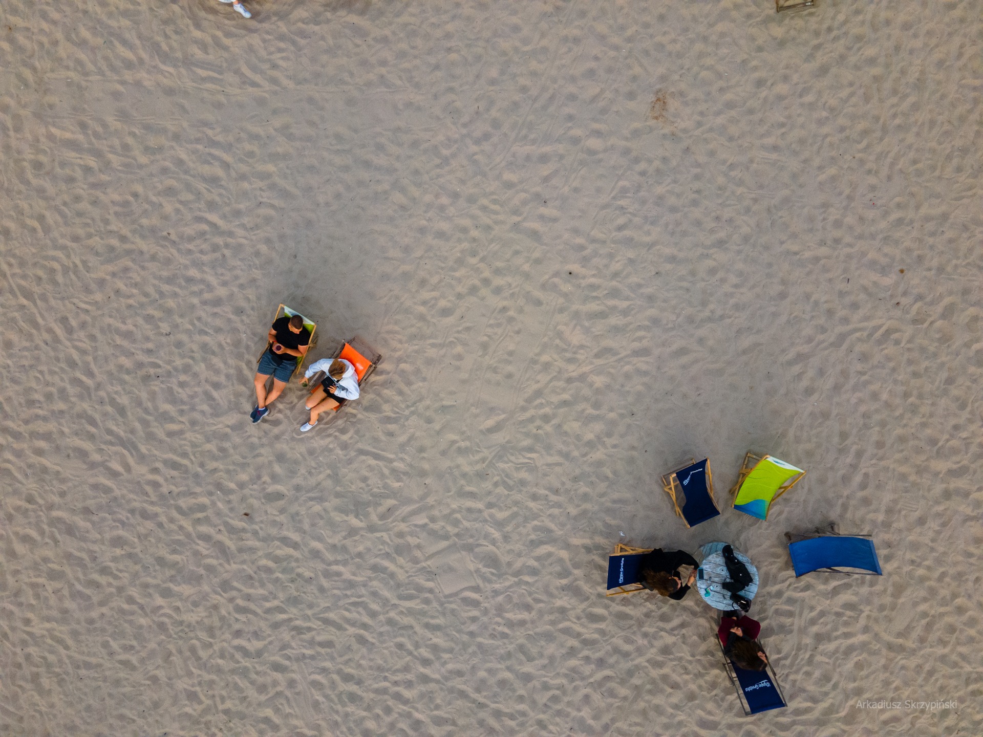 Widok z góry na grupę osób relaksujących się na leżakach na piaszczystej plaży, tworzących kompozycję z wyraźną przestrzenią negatywną.