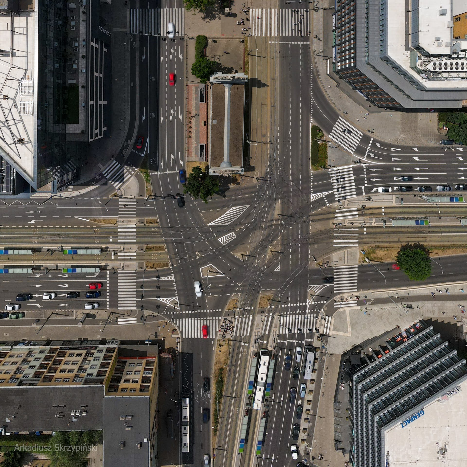 Widok z lotu ptaka na skrzyżowanie z tramwajami, samochodami i pieszymi w słoneczny dzień, z widocznymi budynkami po bokach.