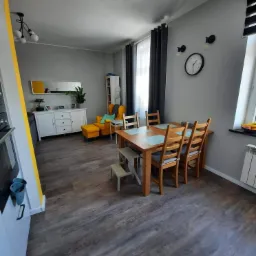 Szare wnętrze pokoju dziennego z drewnianą podłogą, stołem i krzesłami, żółtym fotelem i komodą, oraz dekoracyjnymi lampami na ścianie.