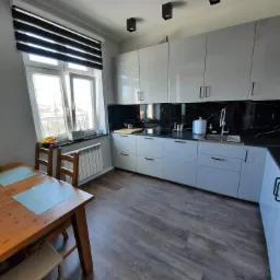 Nowoczesna kuchnia z białymi, lakierowanymi szafkami, blatem i backsplashiem z czarnego marmuru, drewnianym stołem jadalnym i żaluzjami dzień-noc w oknie.