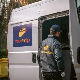 Pracownik w kurtce z logo firmy 'moveandgo' stoi przy otwartych drzwiach białego busa z naklejonym logo firmy, widoczny budynek z czerwonej cegły w tle.
