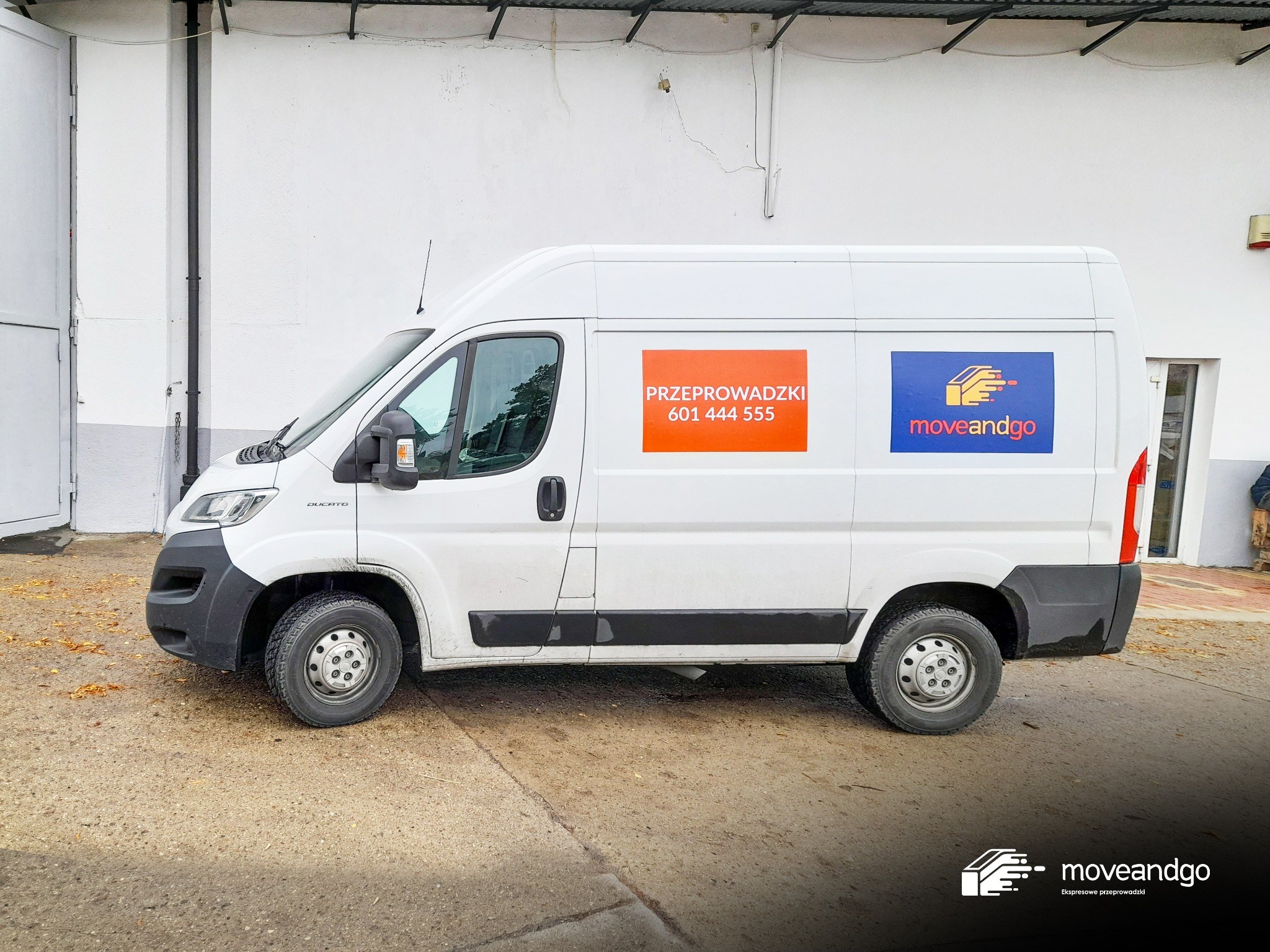 Biały bus dostawczy z naklejkami reklamowymi, w tym pomarańczowa z napisem 'Przeprowadzki' i numerem telefonu, oraz niebieska z logo firmy 'moveandgo', zaparkowany przed białą ścianą budynku.