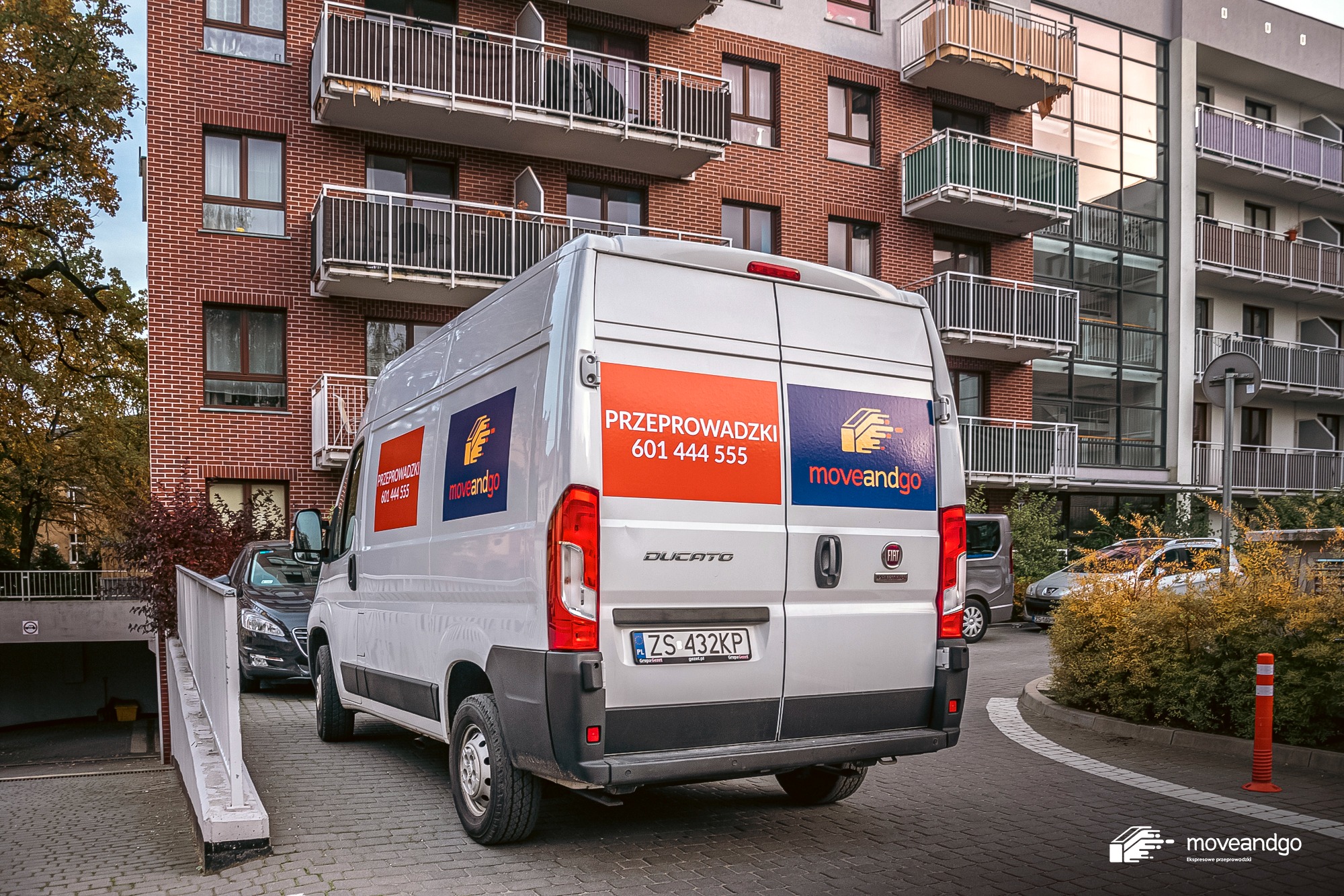 Biały samochód dostawczy marki Fiat Ducato z naklejkami firmy przeprowadzkowej 'moveandgo' zaparkowany na brukowanej ulicy przed nowoczesnym budynkiem mieszkalnym z balkonami.