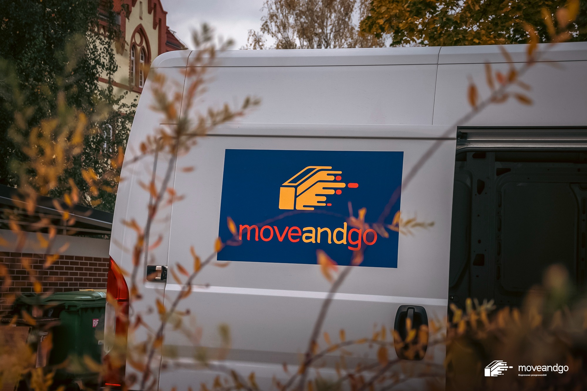 Biały samochód dostawczy z otwartymi drzwiami, z logo firmy 'moveandgo' w Szczecinie, widoczne jesienne liście na pierwszym planie i budynek w tle.