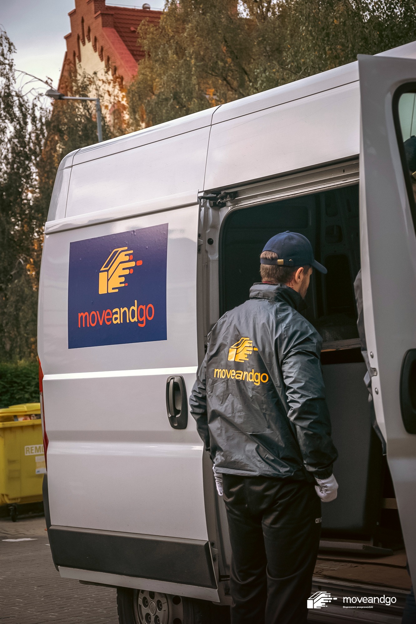 Pracownik w kurtce z logo firmy 'moveandgo' stoi przy otwartych drzwiach białego busa z naklejonym logo firmy, widoczny budynek z czerwonej cegły w tle.