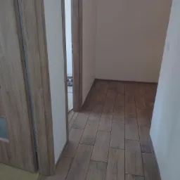 Korytarz z nową podłogą z płytek imitujących drewno, widoczna ościeżnica i fragment drzwi z szybką, w tle puste pomieszczenie z identyczną podłogą.