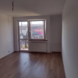 Pusty pokój po remoncie z ułożonym parkietem w odcieniach brązu, białymi ścianami, oknem z widokiem na ulicę i wiszącą żarówką bez klosza.