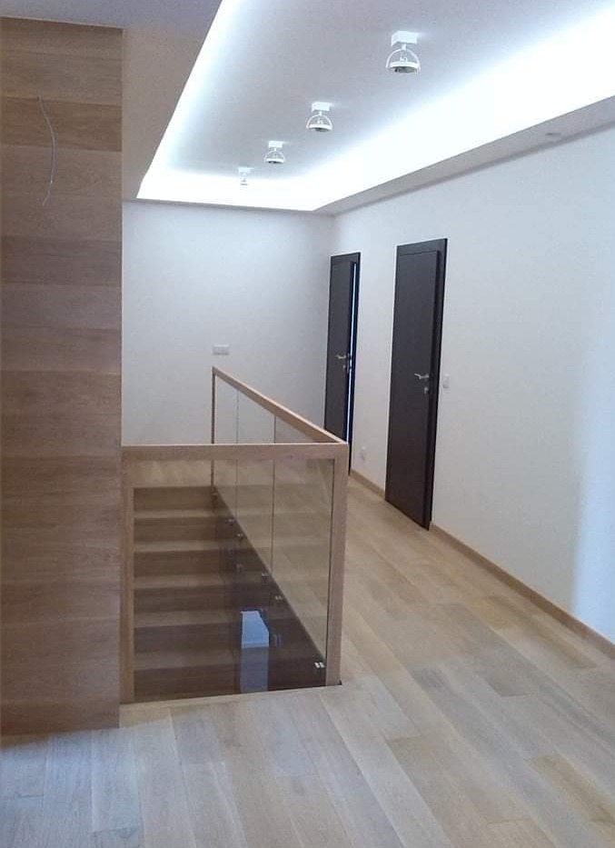 Nowoczesne wnętrze z drewnianą podłogą, szklaną balustradą schodów oraz dwoma ciemnymi drzwiami w białej ścianie, oświetlone punktowymi lampami sufitowymi.