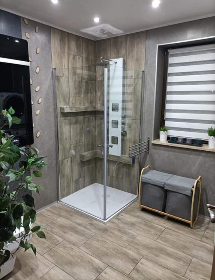 Nowoczesna kabina prysznicowa z hydromasażem w łazience wykończonej płytkami imitującymi drewno, z widoczną rośliną doniczkową i pojemnikiem na pranie.