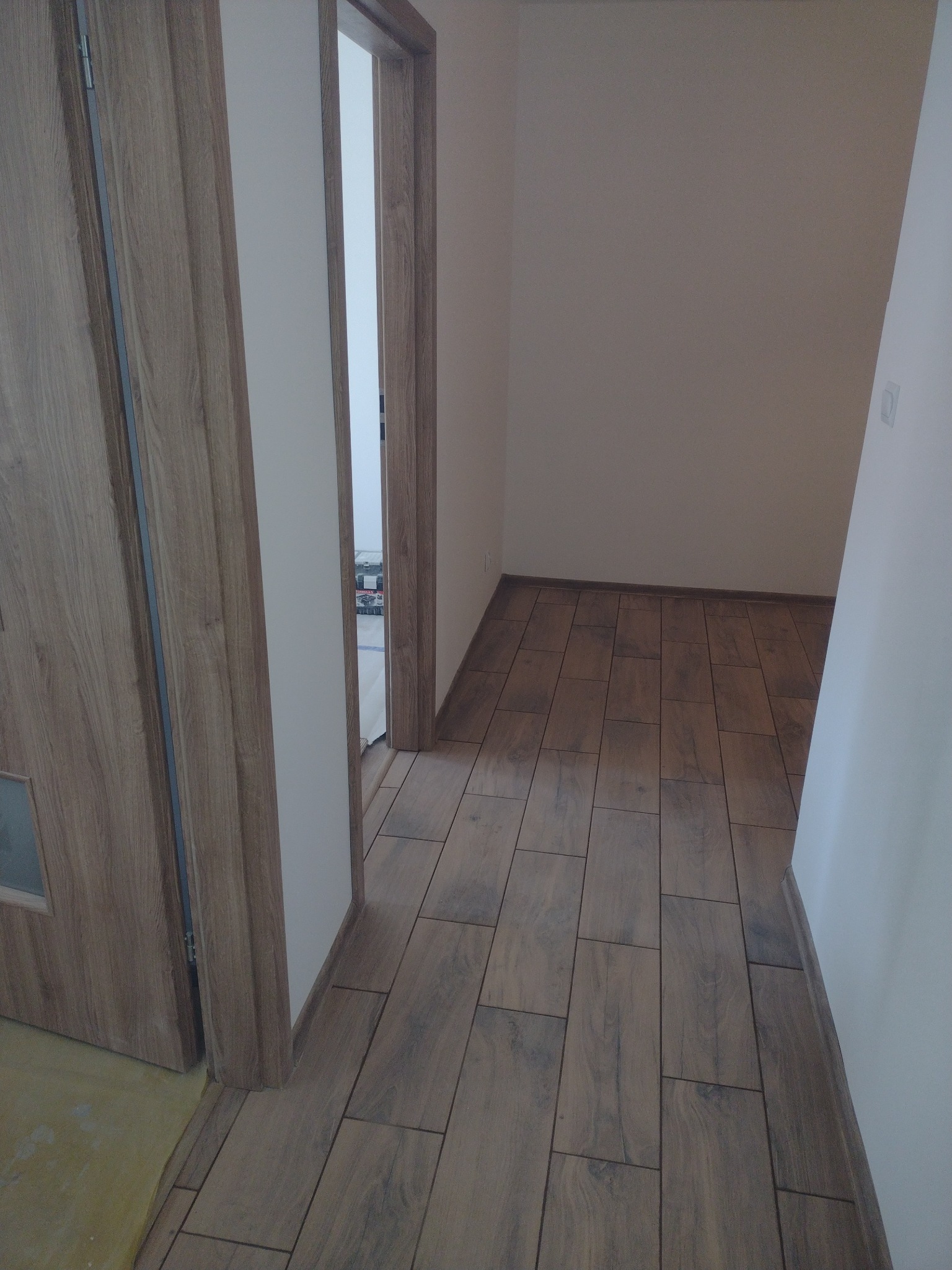 Korytarz z nową podłogą z płytek imitujących drewno, widoczna ościeżnica i fragment drzwi z szybką, w tle puste pomieszczenie z identyczną podłogą.