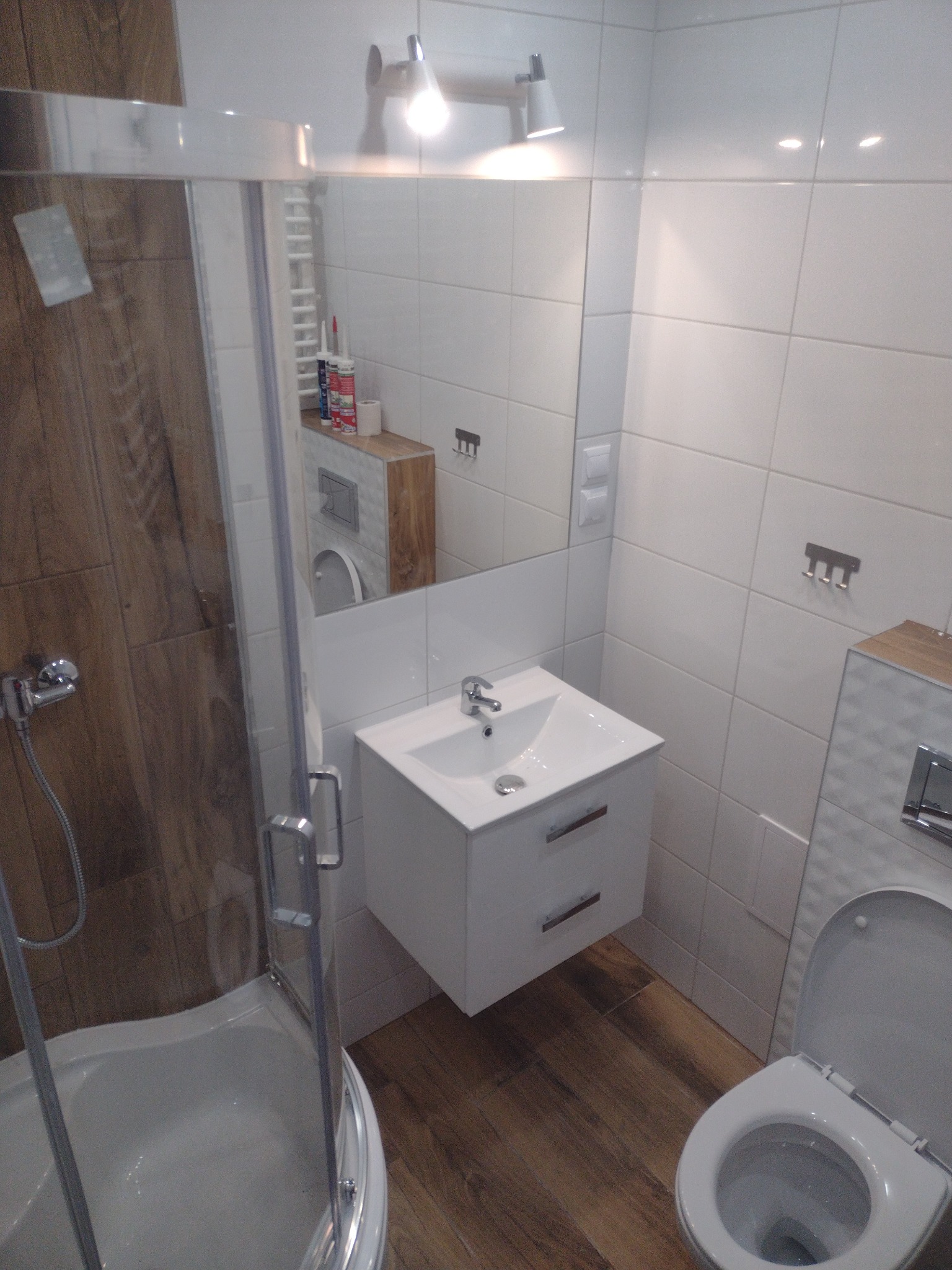 Nowoczesna łazienka z kabiną prysznicową z drewnopodobnymi płytkami, białą umywalką z szafką, toaletą i lustrem z oświetleniem punktowym.