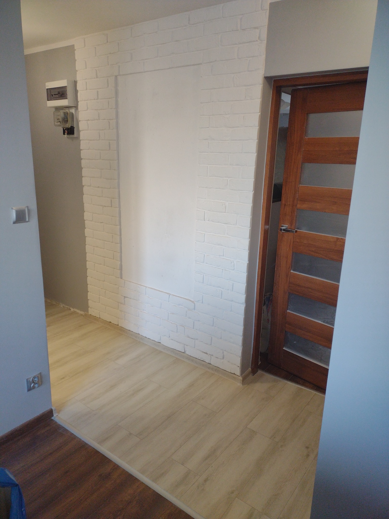 Korytarz z drewnianymi panelami podłogowymi, ścianą z białej cegły z wnęką i drewnianymi drzwiami z przeszkleniami.