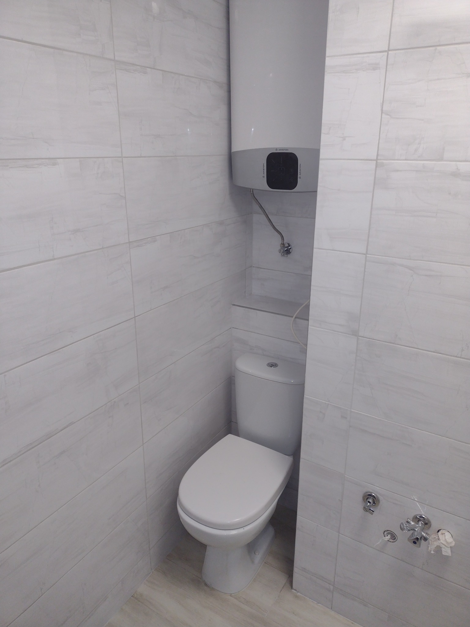 Jasna łazienka z płytkami imitującymi marmur, wiszący bojler Ariston i toaleta.