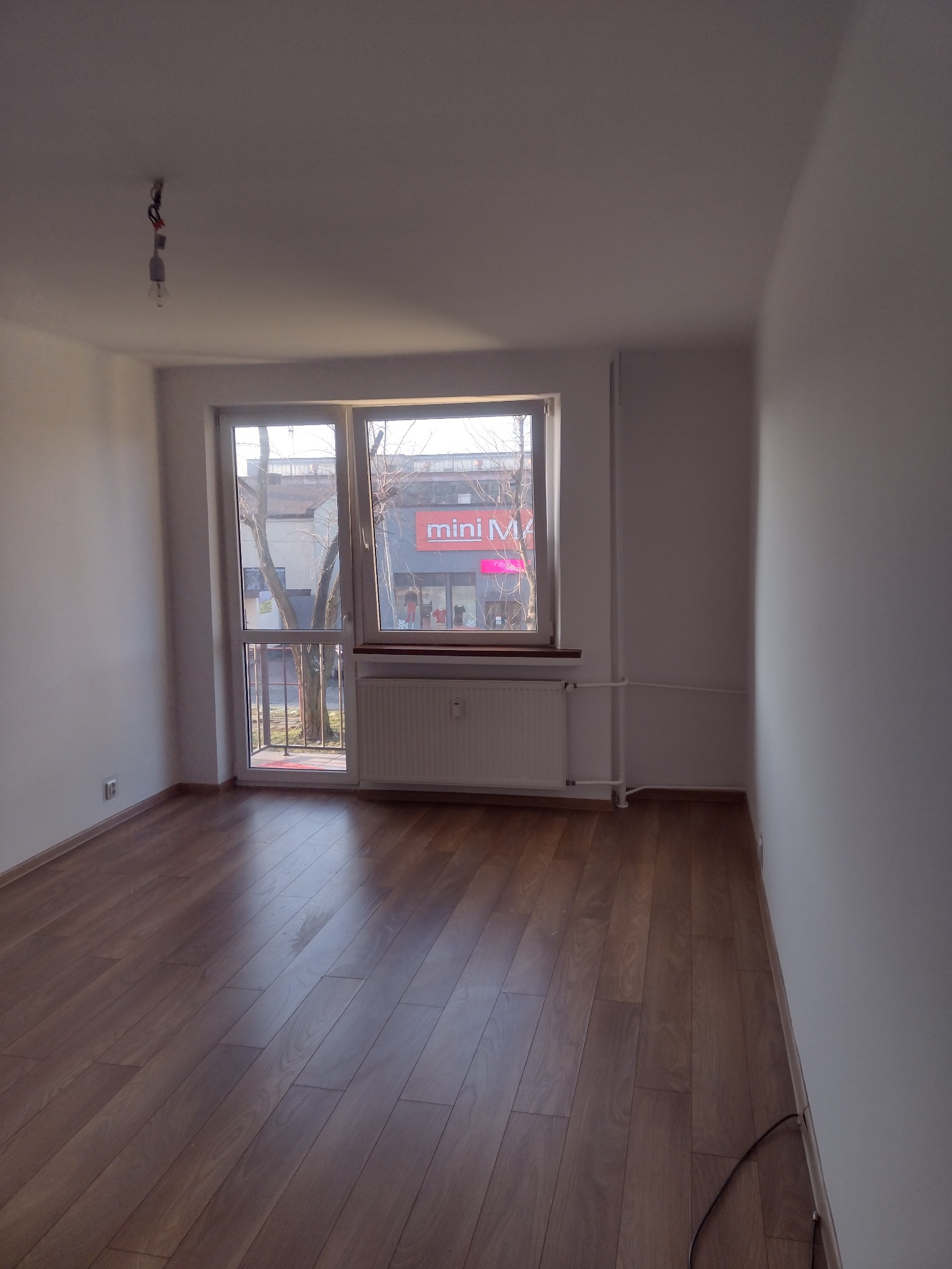 Pusty pokój po remoncie z ułożonym parkietem w odcieniach brązu, białymi ścianami, oknem z widokiem na ulicę i wiszącą żarówką bez klosza.