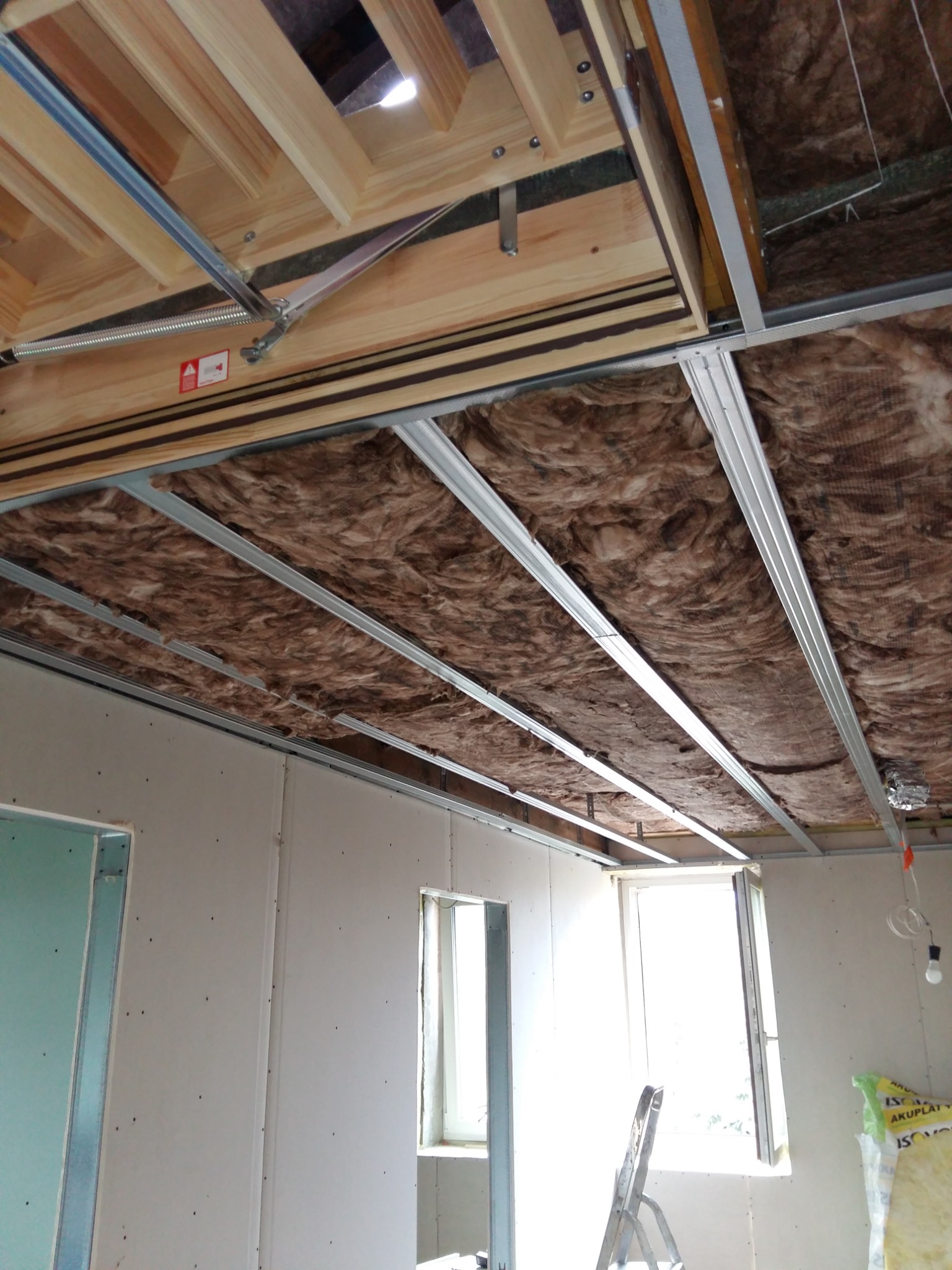 Wnętrze w trakcie prac wykończeniowych: sufit podwieszany z izolacją z wełny mineralnej, widoczne profile metalowe, płyty gipsowo-kartonowe na ścianach z otworami okiennymi, drabina i materiały...