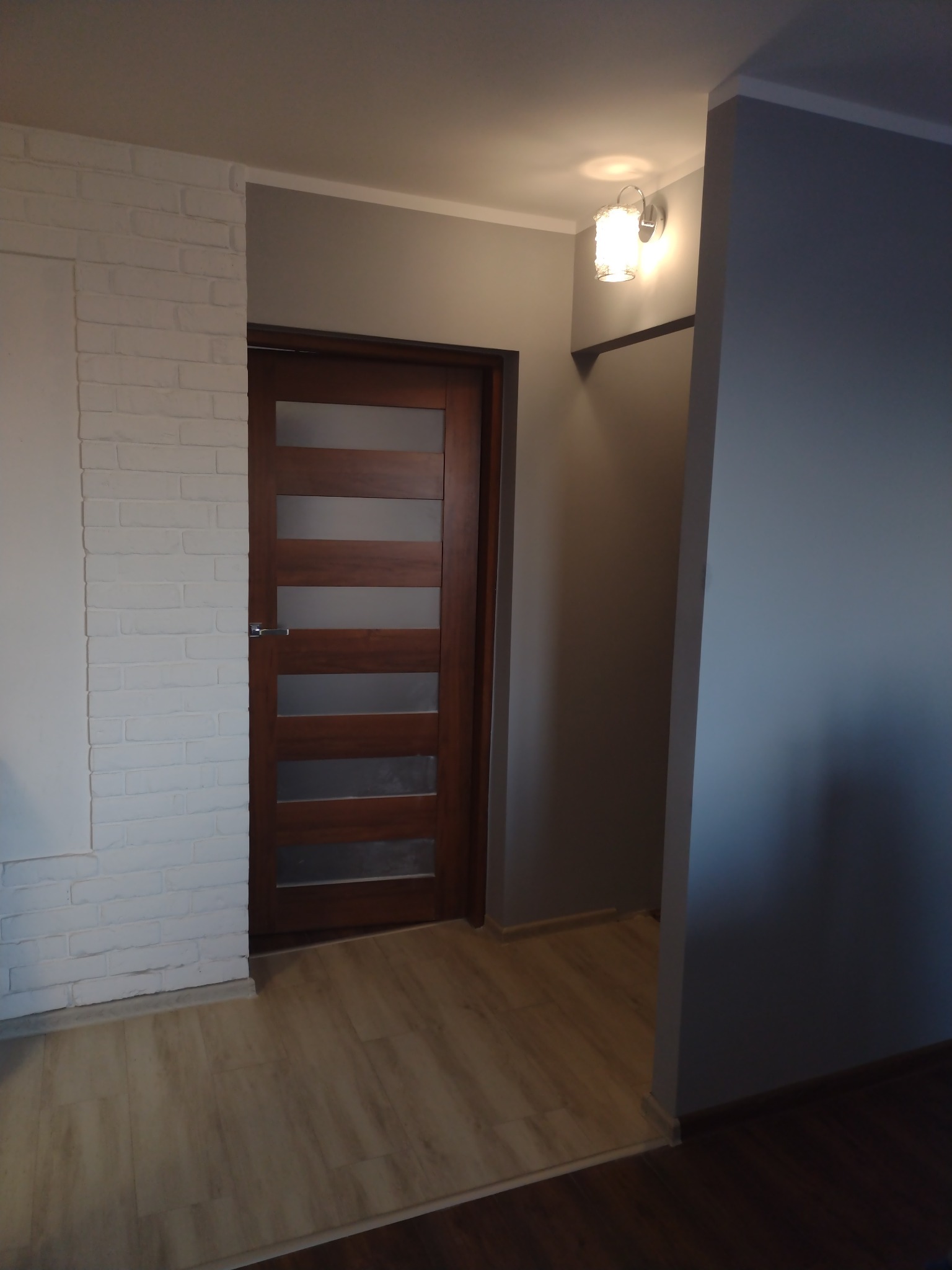 Nowoczesne, brązowe drzwi wewnętrzne z przeszkleniami, zamontowane w odnowionym korytarzu z białą cegłą na ścianie i panelami podłogowymi o jasnym odcieniu drewna.