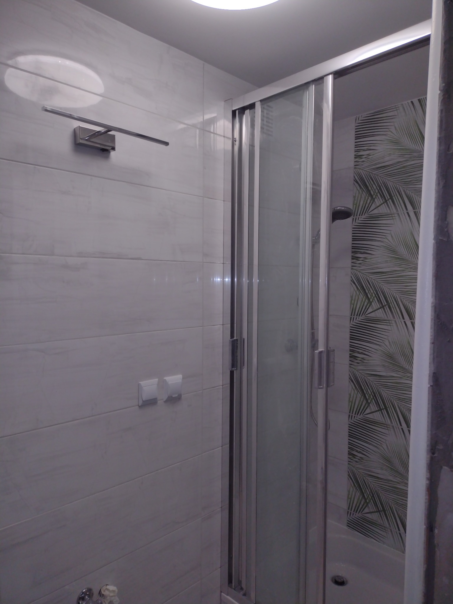 Nowoczesna kabina prysznicowa z przesuwnymi drzwiami i wzorem tropikalnych liści na ścianie wewnątrz, oświetlenie punktowe i chromowana armatura.