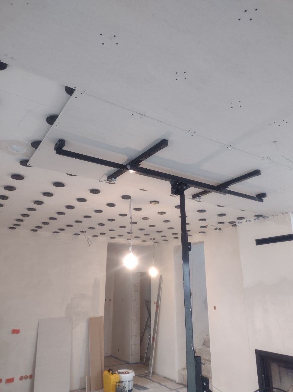 Montaż płyty gipsowo-kartonowej na suficie z wykorzystaniem podnośnika, widoczne otwory na oświetlenie punktowe i surowe ściany w tle.