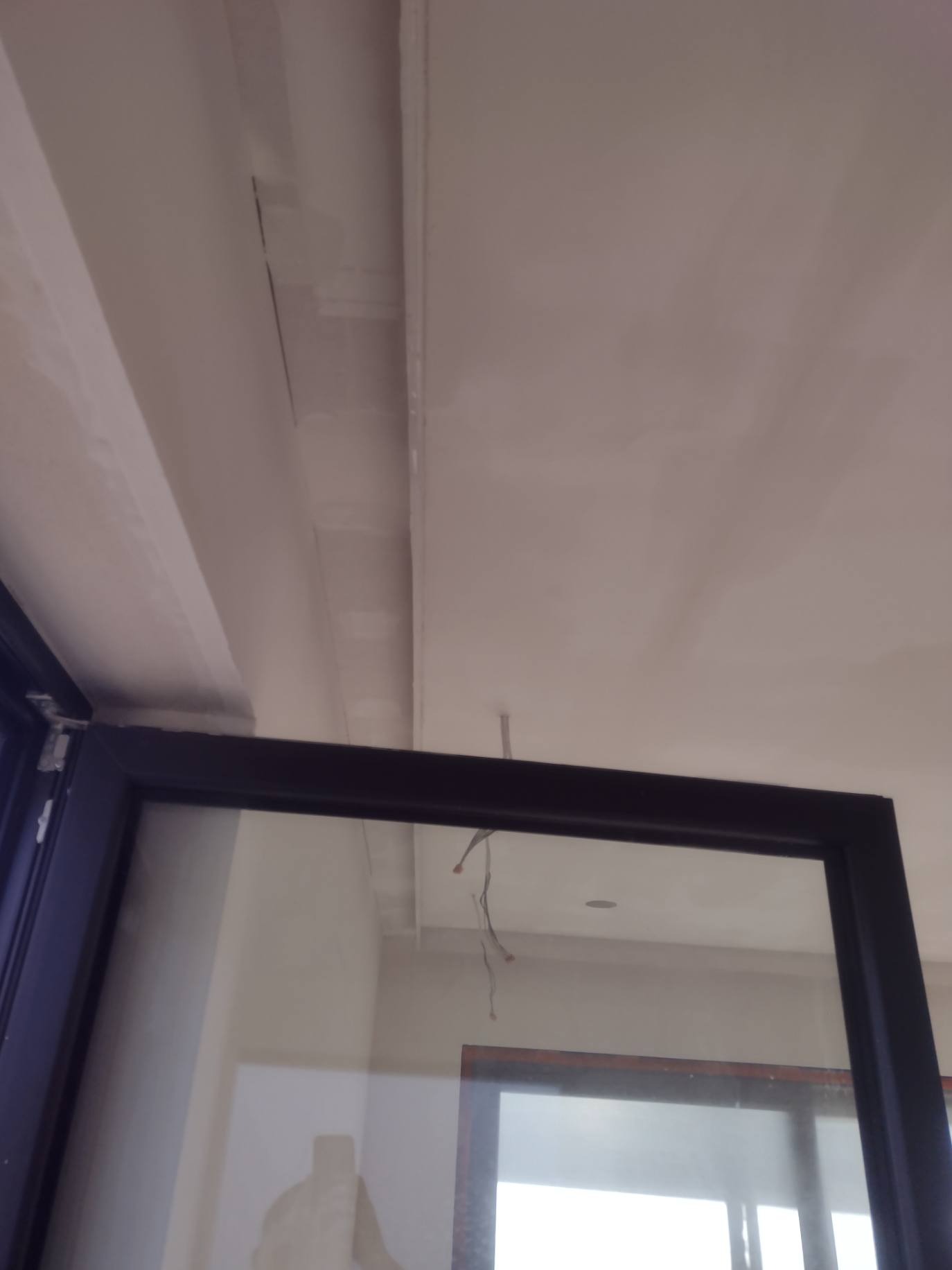 Niedokończona powierzchnia sufitu z widocznymi łączeniami płyt, częściowo zakryta ramą okienną z widocznym odbiciem.