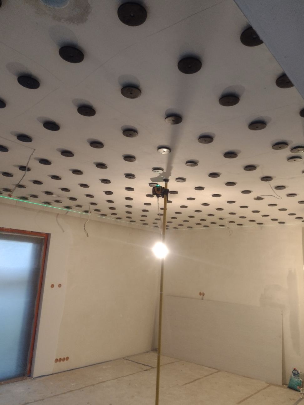 Przygotowany sufit z zamontowanymi elementami do mocowania płyt kartonowo-gipsowych, widoczny laser krzyżowy do poziomowania, ściany przygotowane do dalszych prac wykończeniowych.