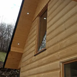 Fragment drewnianego domu z widoczną elewacją z bali, oknami i podbitką dachową z oświetleniem punktowym, na tle błękitnego nieba z chmurami.