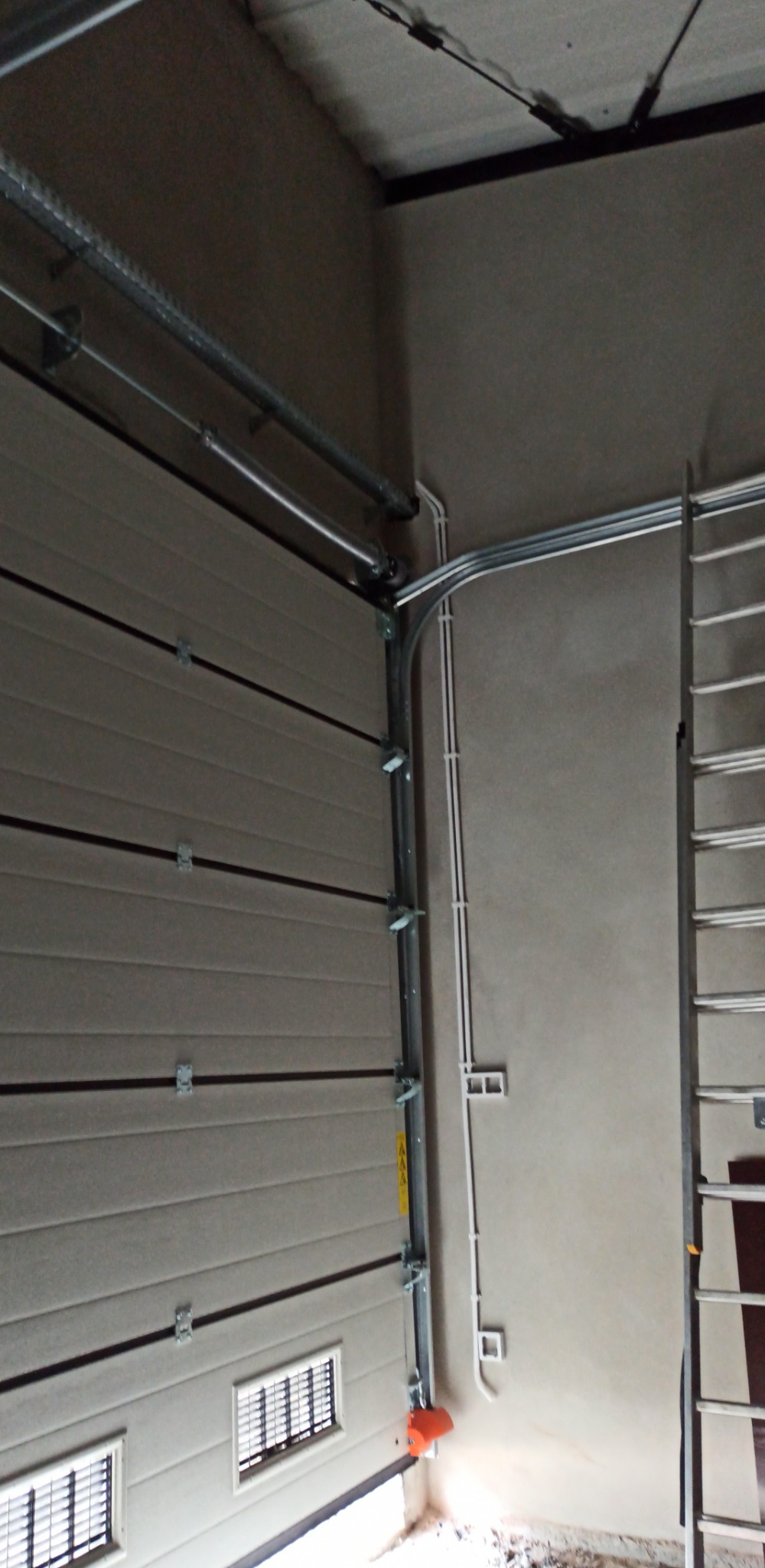 Szara brama garażowa segmentowa z widocznymi elementami mocującymi, prowadnicami i sprężynami, obok ściana z poprowadzonymi przewodami elektrycznymi w białych rurkach, widoczna drabina aluminiowa.