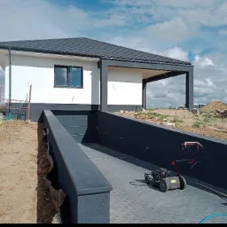 Nowoczesny dom z szarą kostką brukową na podjeździe i kosiarką, widok na elewację w trakcie wykończenia.