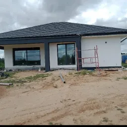 Nowoczesny dom jednorodzinny z białą elewacją i szarymi ramami okiennymi, widoczne rusztowanie przy ścianie bocznej, teren w trakcie przygotowania pod zagospodarowanie.