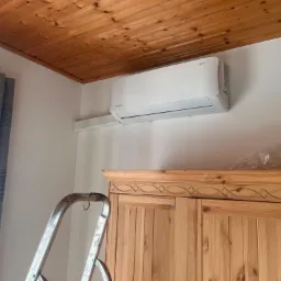 Biały klimatyzator Rotenso zamontowany na ścianie w pomieszczeniu z drewnianym sufitem i szafą, widoczna drabina.