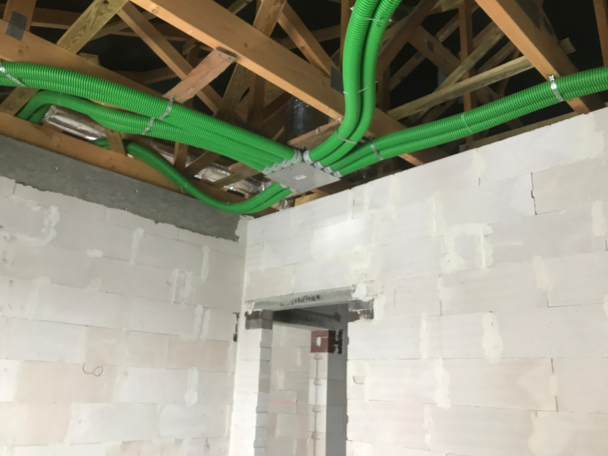 Zielone, karbowane rury wentylacyjne przymocowane do drewnianej konstrukcji dachu w surowym wnętrzu z białymi ścianami z betonu komórkowego, widoczne złącze rozdzielające rury.