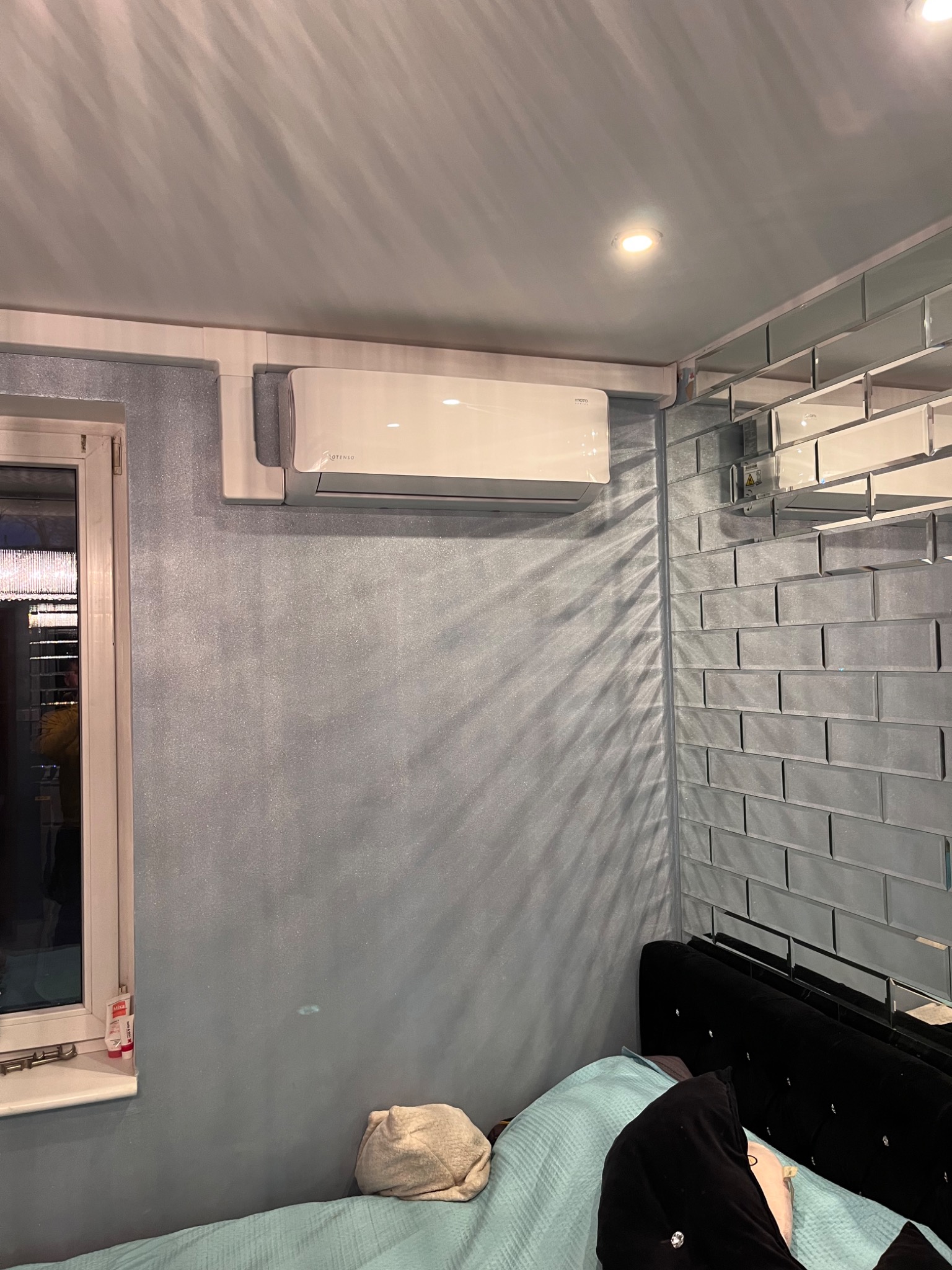 Biały klimatyzator ścienny marki OtensO zamontowany w sypialni z niebieską, błyszczącą ścianą i lustrzaną mozaiką, widoczne łóżko z czarnym zagłówkiem.