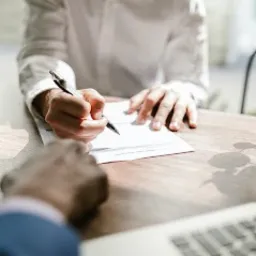 Osoba podpisuje dokument na drewnianym biurku, widoczny fragment laptopa i dłoń drugiej osoby.