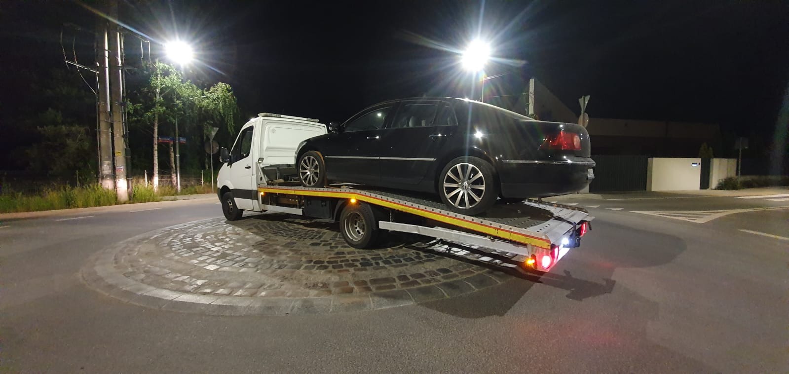 Czarny samochód osobowy marki Volkswagen Phaeton transportowany na żółtej lawecie na tle nocnego krajobrazu miejskiego z oświetlonym rondem.