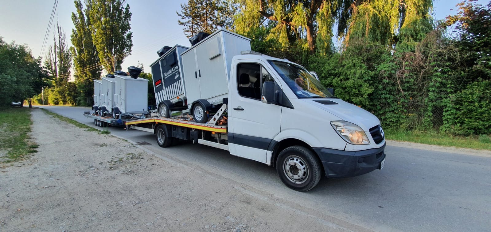 Biała laweta Mercedes-Benz z dwoma przyczepami z kontenerami MobileCam na platformie, widok z boku na tle zieleni.