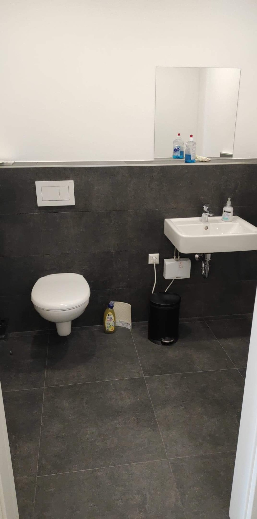 Nowoczesna łazienka z ciemnymi płytkami, wiszącą toaletą i umywalką, minimalistycznym lustrem i akcesoriami do sprzątania na podłodze.