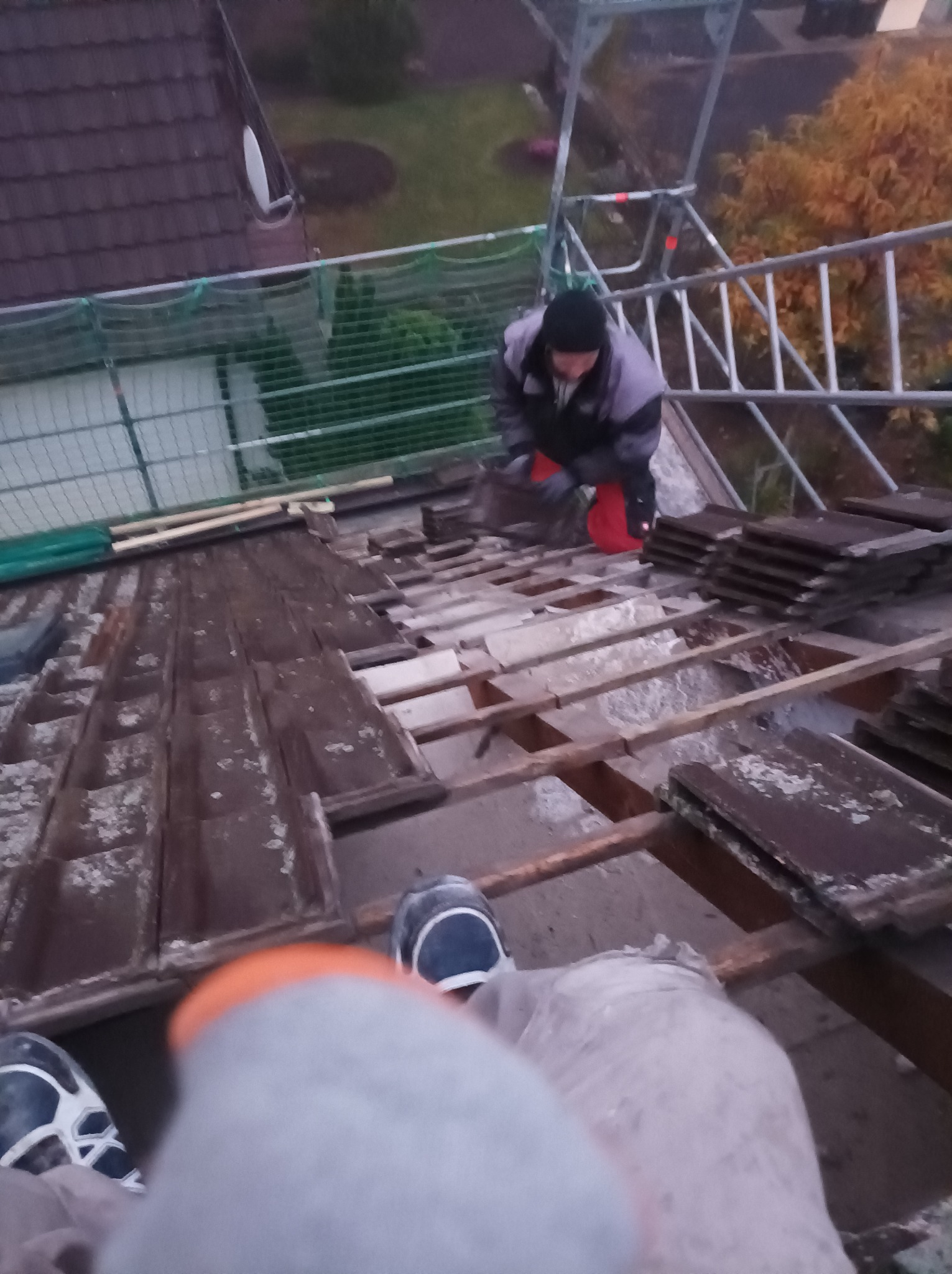 Pracownik w stroju roboczym wymienia stare dachówki na konstrukcji dachu, widoczne rusztowanie i sąsiedni budynek.