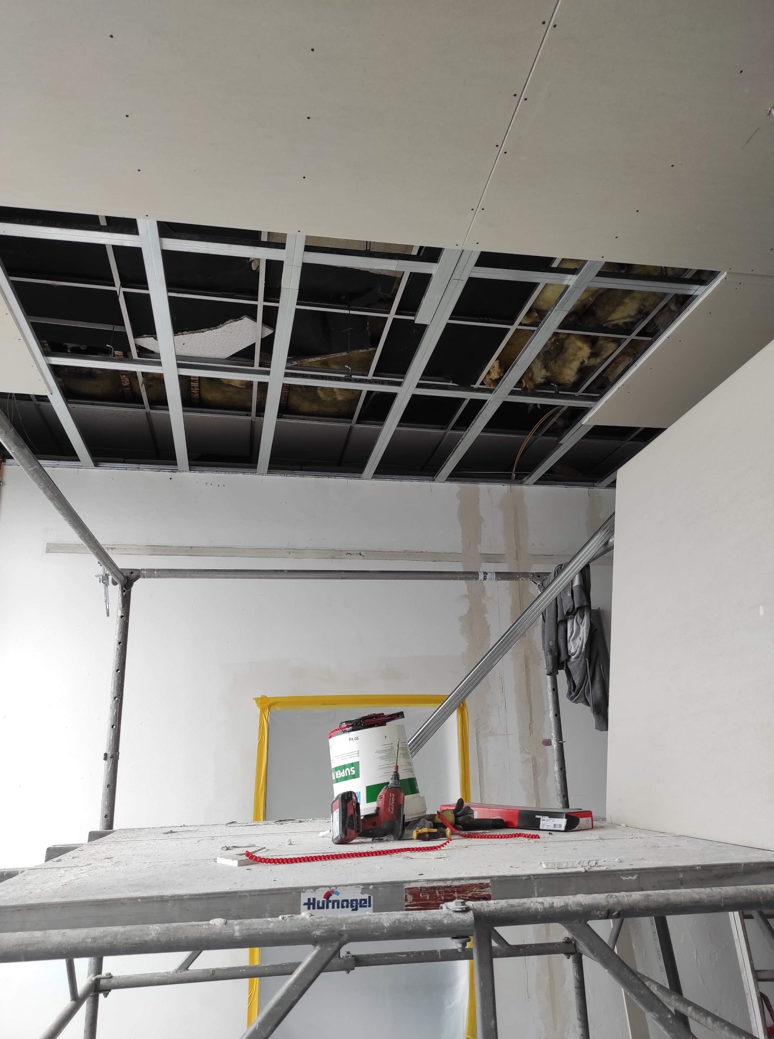 Montaż sufitu podwieszanego z płyty karton gips: widoczne metalowe profile, częściowo ułożona płyta, izolacja oraz narzędzia na rusztowaniu.