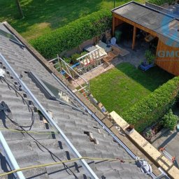Usługi budowlane - Montaż paneli słonecznych na dachu z szarej dachówki, widok z góry na zielony ogród i mały budynek gospodarczy. Widoczne aluminiowe profile i kable.