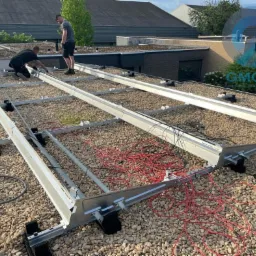 Montaż stelażu pod panele fotowoltaiczne na płaskim dachu pokrytym drobnym żwirem. Widoczne przewody elektryczne i pracownicy w trakcie instalacji.