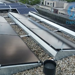 Usługi budowlane - Instalacja paneli słonecznych na płaskim dachu pokrytym żwirem. Widoczne metalowe obudowy paneli i elementy montażowe. W tle budynki.