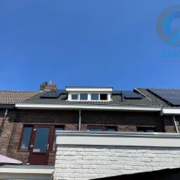 Ceglany dom z panelami słonecznymi na dachu, widok z dołu. Biała, ceglana dobudówka. Błękitne niebo w tle, logo firmy w prawym górnym rogu.