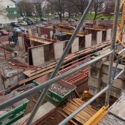 Usługi budowlane - Budowa w toku: widok z góry na szalunki, zbrojenia i kontenery z gruzem, otoczone rusztowaniami. W tle budynki mieszkalne i drzewa. Prace ziemne w trakcie.